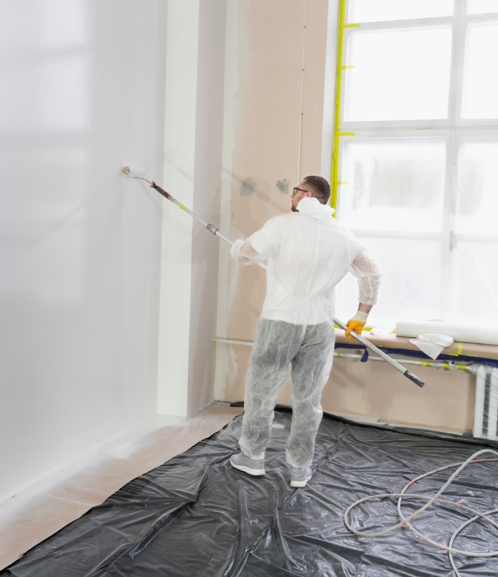 Artisan peintre utilisant un rouleau télescopique pour peindre un mur intérieur protégé par une bâche.